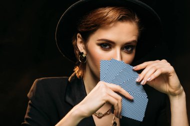 çekici kız kameraya ceket ve siyah, izole poker kartları ile yüz kapsayan şapka