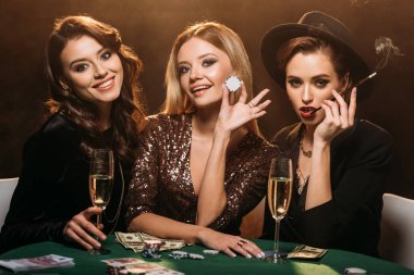 smiling attractive girls sitting at table in casino and looking at camera clipart