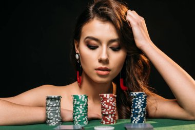 beautiful brown haired girl leaning on table and looking at poker chips isolated on black clipart