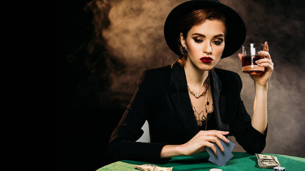 thoughtful attractive girl in jacket and hat playing poker and holding glass of whiskey in casino