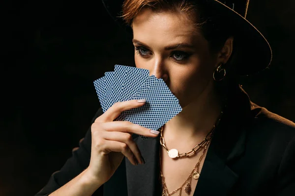 attractive girl in jacket and hat covering face with poker cards isolated on black and looking away