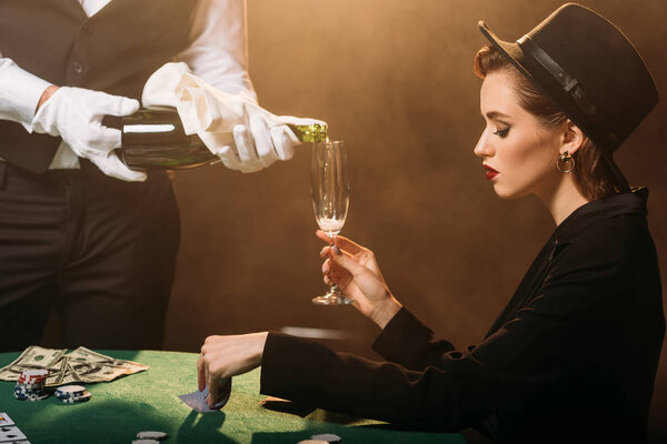 официант наливает шампанское в бокал привлекательной девушки в куртке и шляпе, пока она играет в покер в казино
