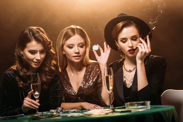 привлекательные девушки с бокалом шампанского, сигареты и фишки для покера сидя за столом и глядя на покер карты в казино
