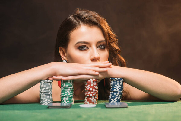beautiful brown haired girl leaning on poker chips and looking at camera isolated on black