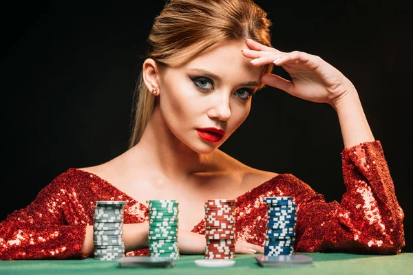 attractive girl in red sparkling dress leaning on table with poker chips isolated on black