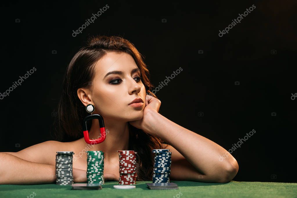 Beautiful brown haired girl resting chin on hand and looking away, poker chips on table isolated on black