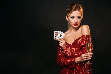 attractive girl in red shiny dress holding joker and queen of hearts cards isolated on black, looking at camera clipart