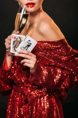 cropped image of girl in red shiny dress holding joker and queen of hearts cards, drinking champagne isolated on black clipart