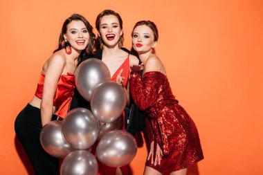 laughing beautiful girls in stylish party clothes holding bundle of grey balloons and looking at camera isolated on orange clipart