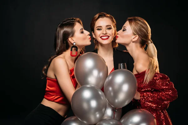 two girls in stylish party clothes kissing friend, she holding bundle of grey balloons isolated on black