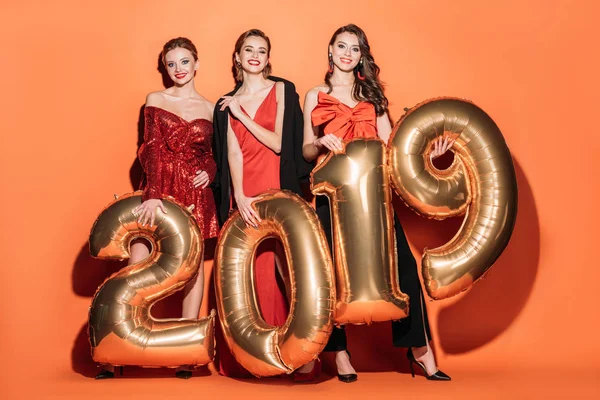 smiling attractive girls in stylish party clothes holding 2019 and looking at camera balloons on orange