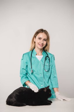 cheerful female veterinarian standing near black cat on grey background clipart