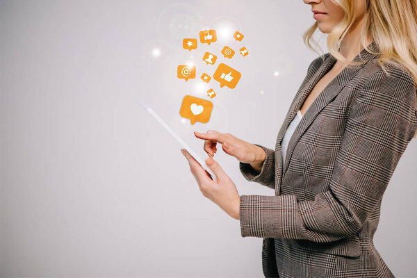 cropped view on businesswoman in suit touching digital tablet with multimedia icons isolated on grey