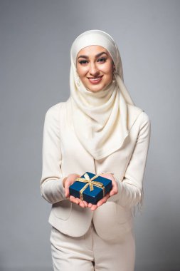 happy young muslim woman holding gift box and smiling at camera isolated on grey clipart