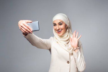 mutlu genç Müslüman kadın gri izole smartphone ile selfie alarak 