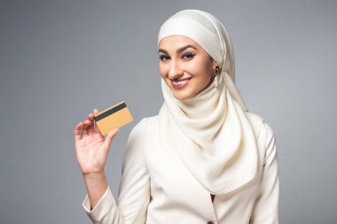 kredi kartı tutan ve gri izole kamera gülümseyen genç Müslüman kadın 