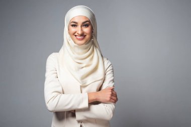 çapraz kolları ile ayakta ve gri izole kamera gülümseyen genç Müslüman kadın