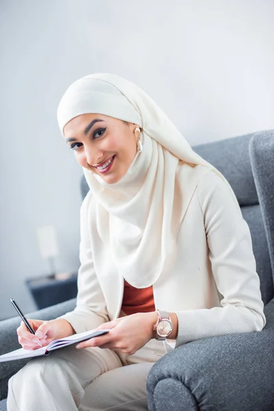 年轻的穆斯林妇女写在笔记本和微笑在相机 — 图库照片
