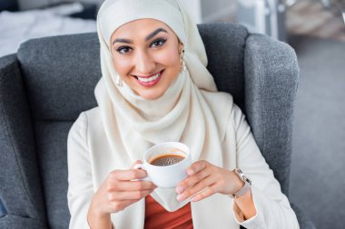 kahve tutarak ve kameraya gülümseyen genç Müslüman kadın