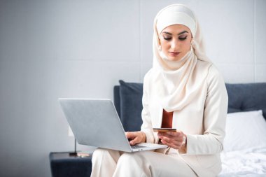 kredi kartı tutan ve dizüstü bilgisayar kullanarak genç Müslüman kadın 