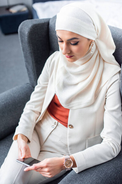 beautiful young muslim woman sitting in armchair and using smartphone 