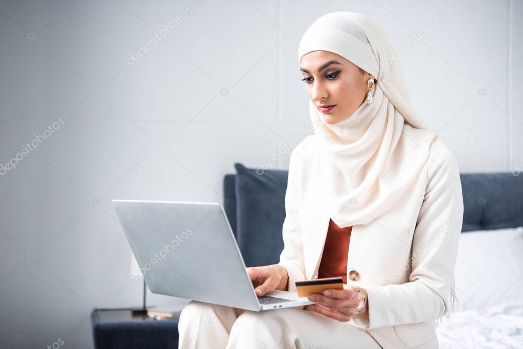 young muslim woman holding credit card and using laptop at home