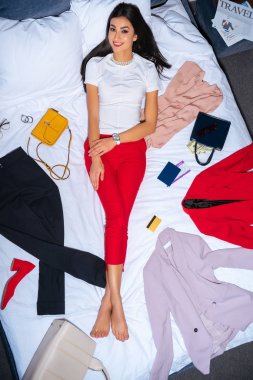 moda giysiler, pasaport, kredi kartı ve seyahat gazetesi ile yatakta yatarken ise kameraya gülümseyen güzel kızın üstten görünüm 