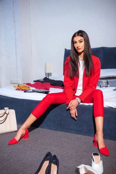 attractive young woman in stylish red suit looking at camera, various fashionable shoes on floor and clothes on bed