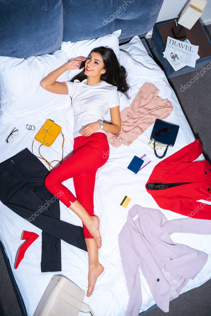 high angle view of beautiful smiling girl talking by smartphone while lying on bed with fashionable clothes, credit card, passport and boarding pass