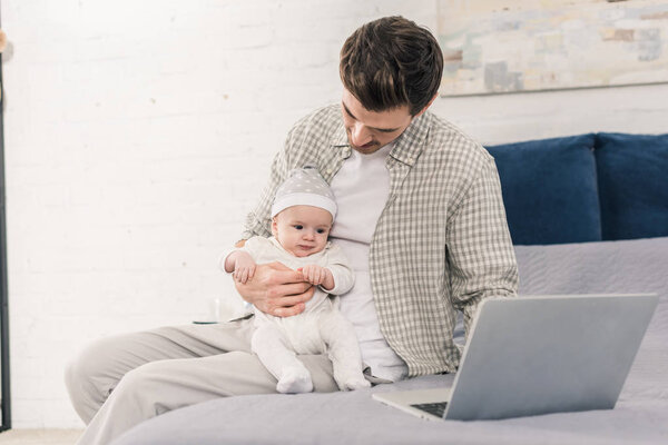 portrait of man working on laptop with little baby in hands at home, work and life balance concept