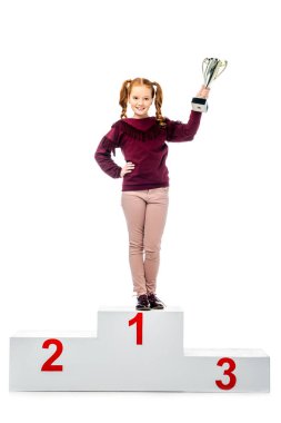 cheerful schoolgirl standing on winner podium, smiling, holding trophy cup and looking at camera isolated on white clipart