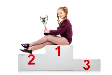 happy schoolgirl sitting on winner podium, holding trophy cup and looking at camera isolated on white clipart