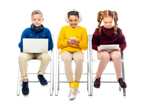 cute children sitting on chairs and using digital devices isolated on white