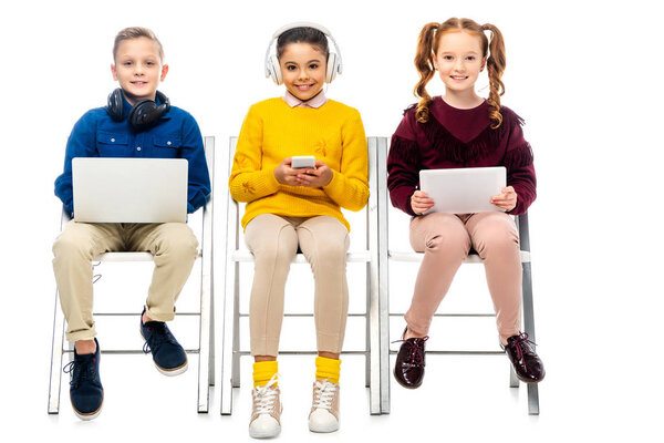 cute kids sitting on chairs, holding digital devices and looking at camera isolated on white