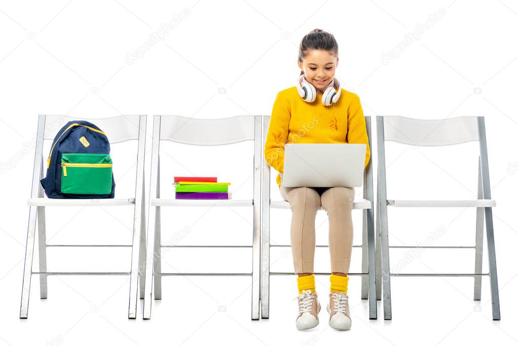 cute schoolgirl sitting on chair and using laptop isolated on white
