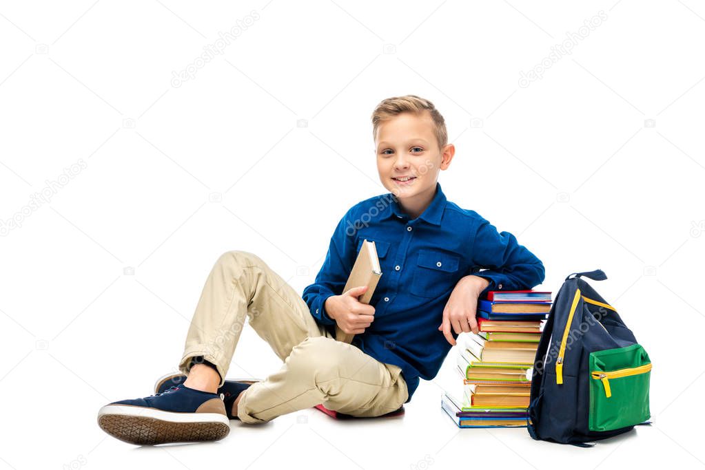 smiling schoolboy holding book, looking at camera and sitting near backpack and stack of books isolated on white