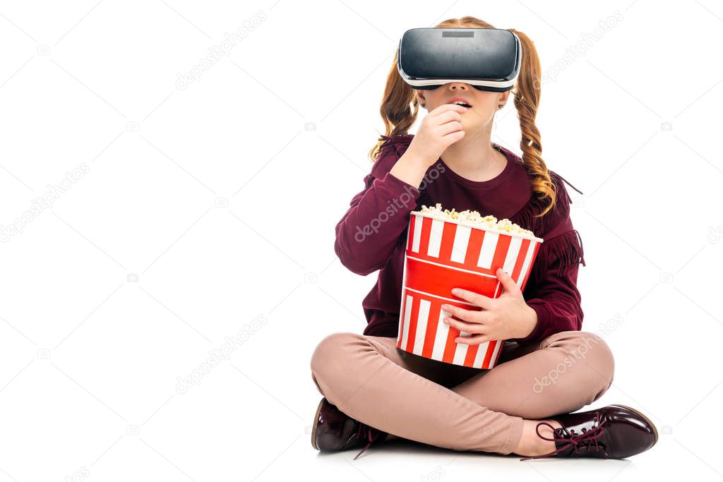 child with virtual reality headset on head holding striped bucket and eating popcorn isolated on white