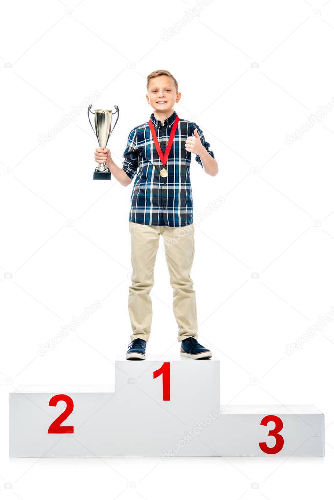 smiling boy standing on winner pedestal, holding trophy cup, showing thumb up and looking at camera isolated on white
