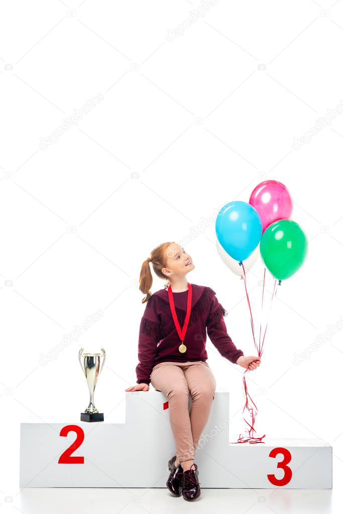 smiling schoolgirl sitting on winner podium near trophy cup and holding colorful balloons isolated on white