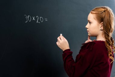thoughtful schoolgirl holding chalk and looking at blackboard with math example clipart