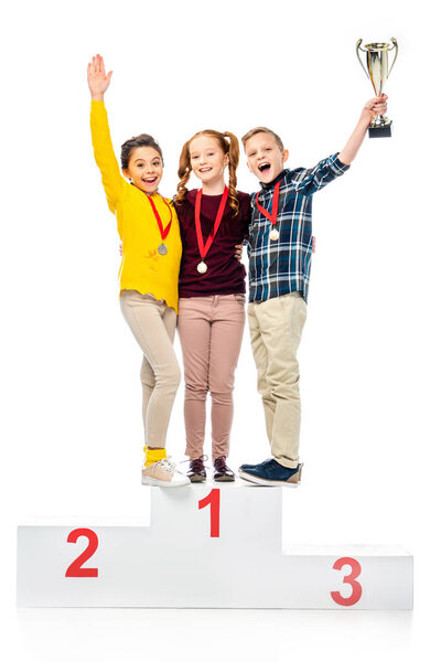 счастливые дети с медалями и кубком трофеев стоя на пьедестале победителя, крича и глядя на камеру изолированы на белом
