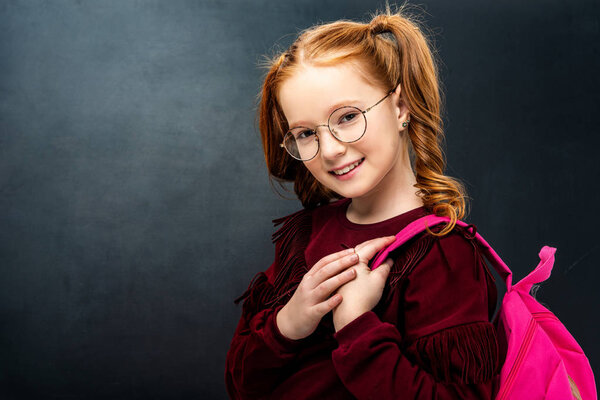 smiling schoolgirl in glasses with pink backpack looking at camera on black background