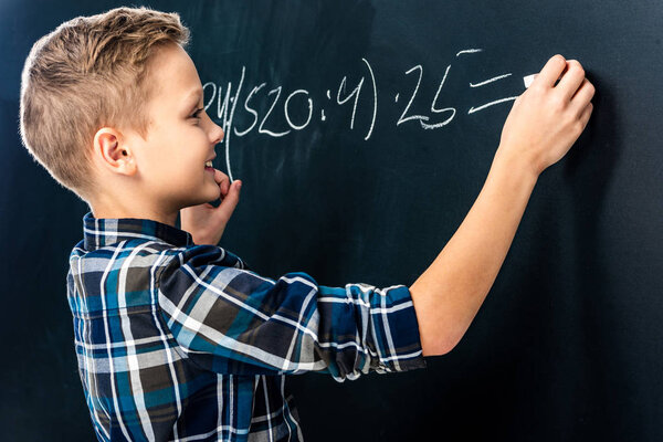 smiling boy writing math example on blackboard with chalk