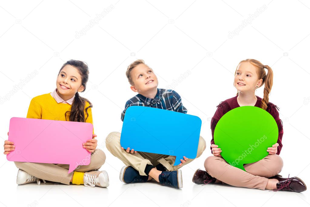 cheerful kids sitting and holding multicolored speech bubbles isolated on white