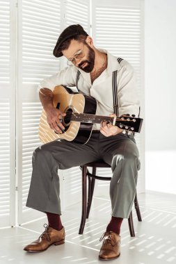 handsome bearded musician in cap sitting on chair and playing acoustic guitar  clipart