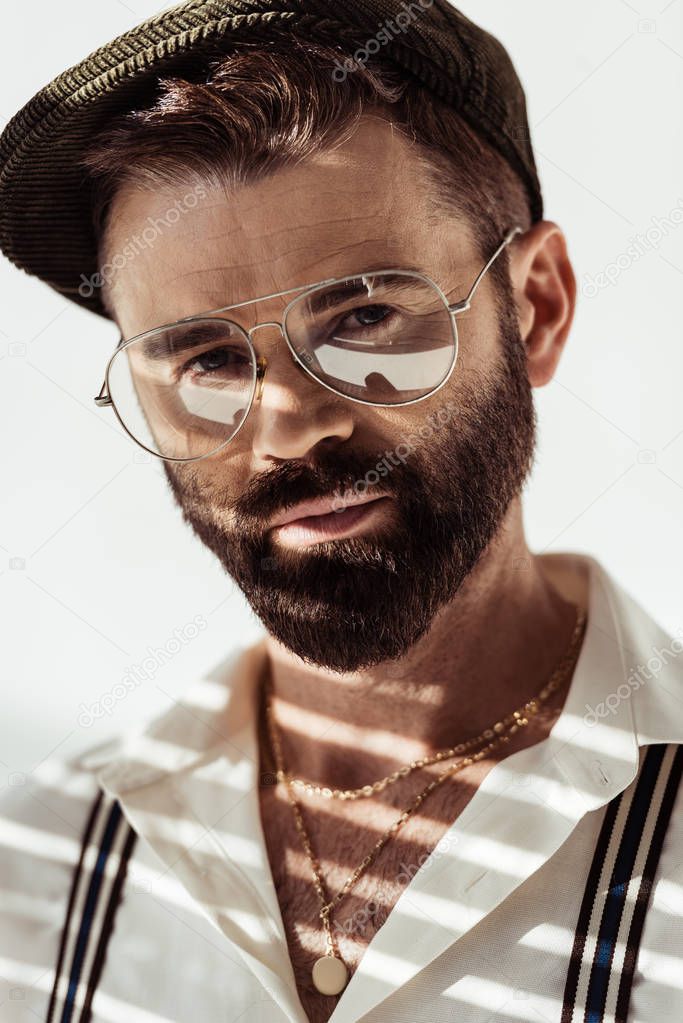 close up view of handsome bearded man in glasses and cap looking at camera on white background