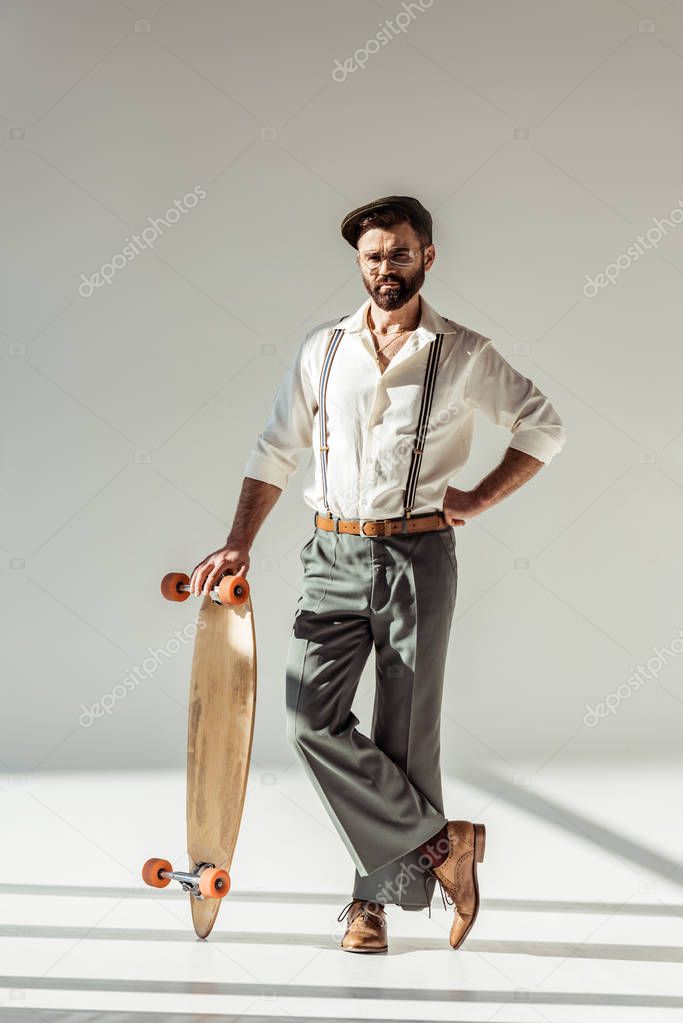 handsome bearded man holding longboard and looking at camera on grey background