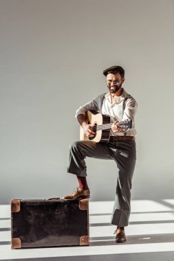 Akustik gitar çalmak ve gri arka plan üzerinde kameraya bakarak vintage çanta yakınındaki şapkalı sakallı adam gülümseyen