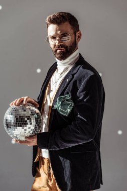 handsome man in glasses holding disco ball and looking at camera on grey background clipart