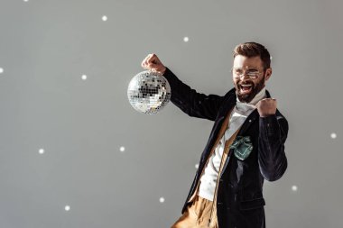 cheerful man in glasses holding disco ball and looking at camera on grey background clipart
